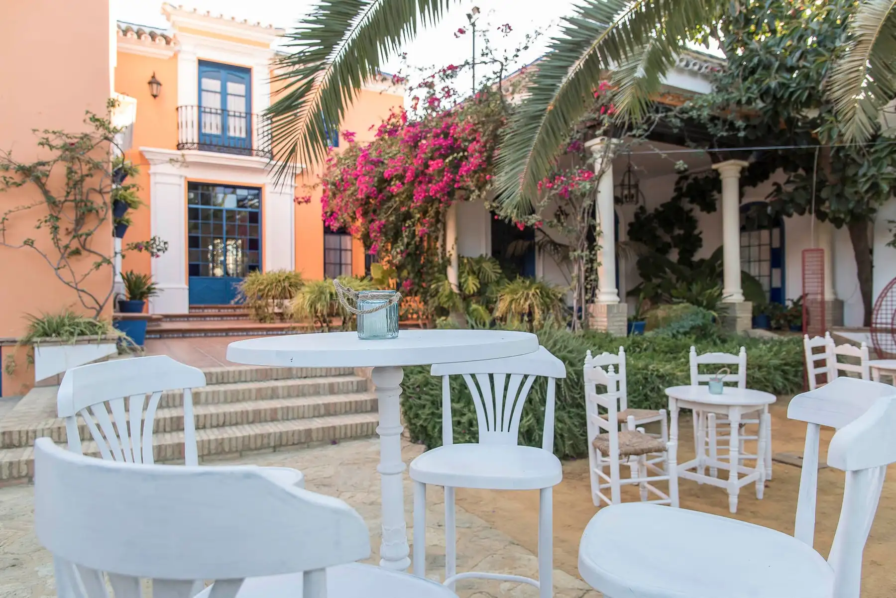 El patio con mesas altas y sillas blancas bajo palmeras y buganvillas en flor, delante de la casa de balcones azules