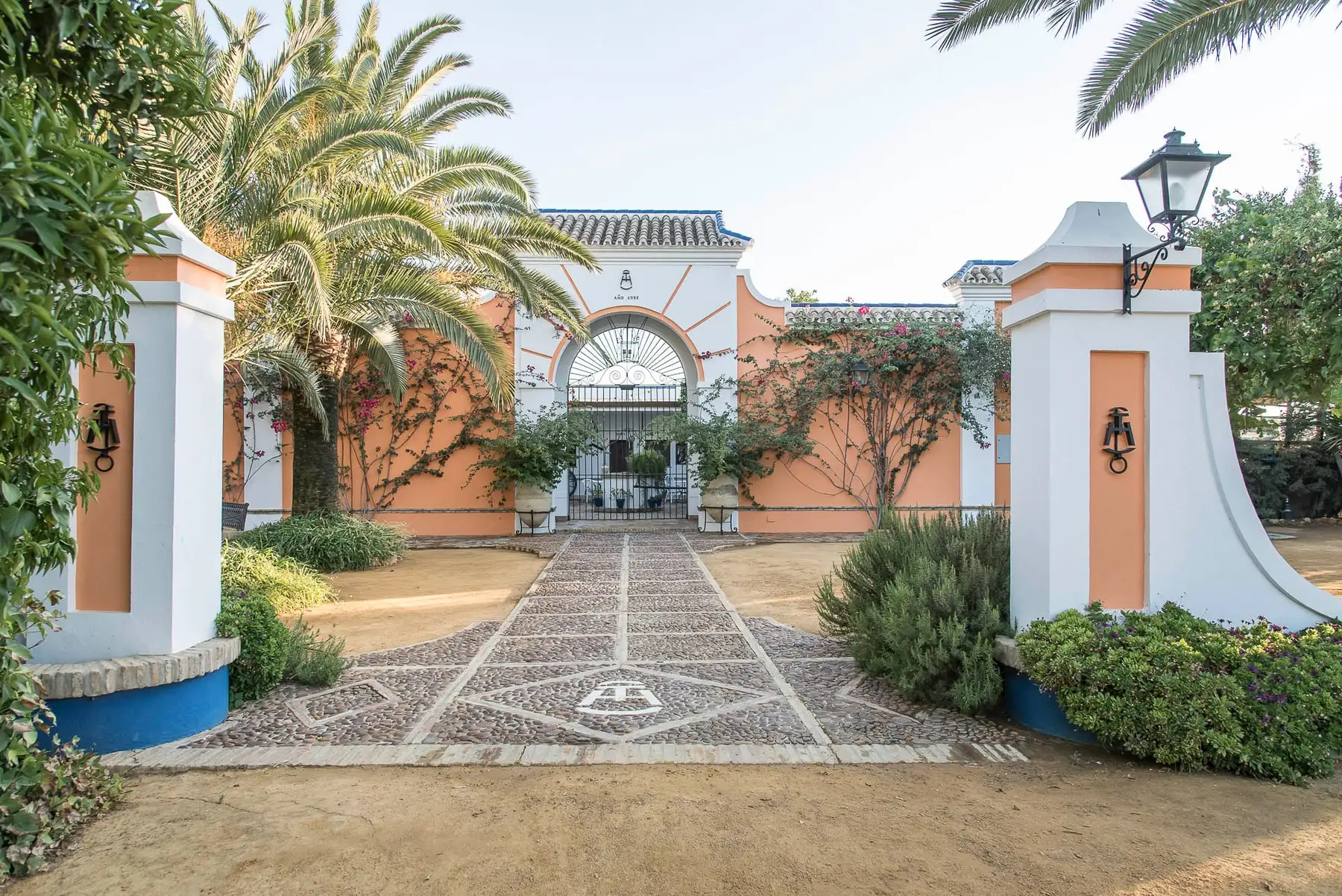 La entrada de la hacienda vista desde el camino de guijarros: pilares blancos con farol, setos, una palmera y el arco al fondo