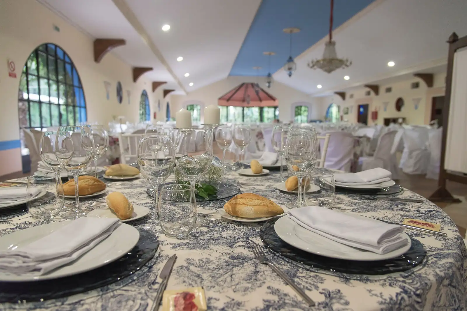 El salón de celebraciones con la mesa puesta: copas, pan y mantel de motivos azules, bajo vigas de madera y un techo azul
